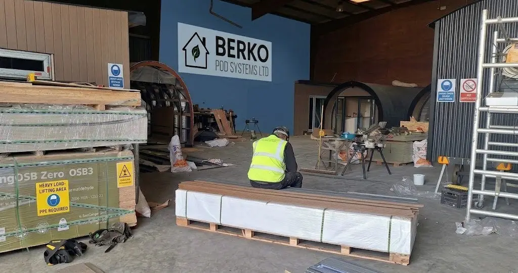 Manufacturing floor of Berko Pod Systems Ltd. featuring modular pod constructions, raw materials, a worker in safety gear, and a prominent company sign.